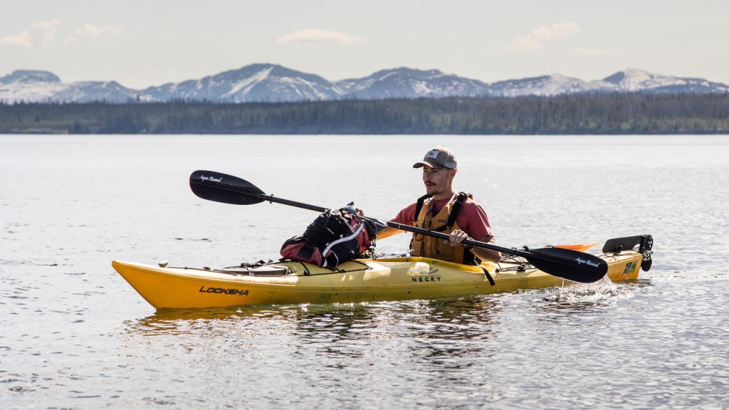 A man in a yellow kayak on a lake with mountains in the background.